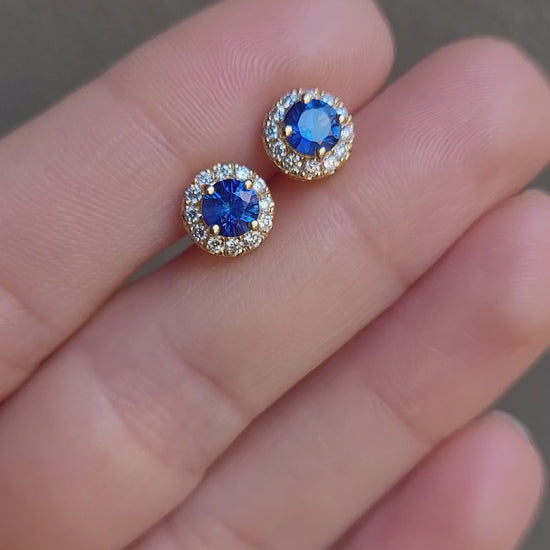 video of Blue Sapphire and Diamond Halo Stud Earrings in 18K Yellow Gold held between fingers in neutral background