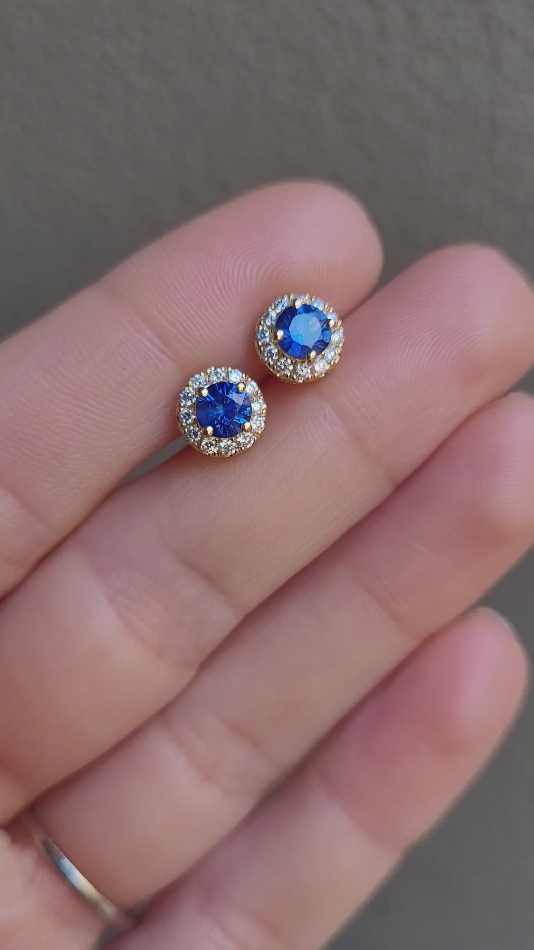 video of Blue Sapphire and Diamond Halo Stud Earrings in 18K Yellow Gold held between fingers in neutral background